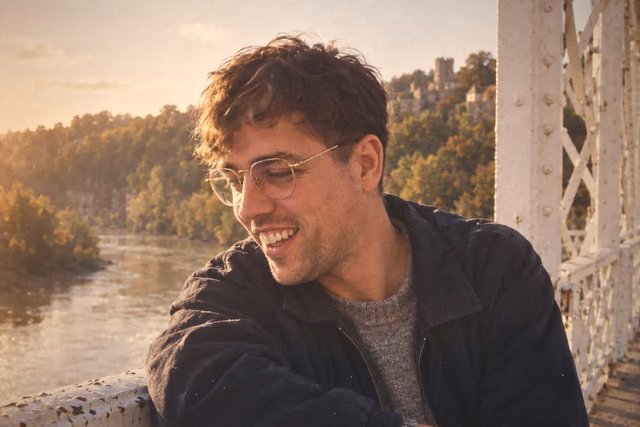 Oliver on the old bridge in Chepstow, laughing and looking away down the river, in glasses and a dark jacket, with the castle on the cliff behind him.
